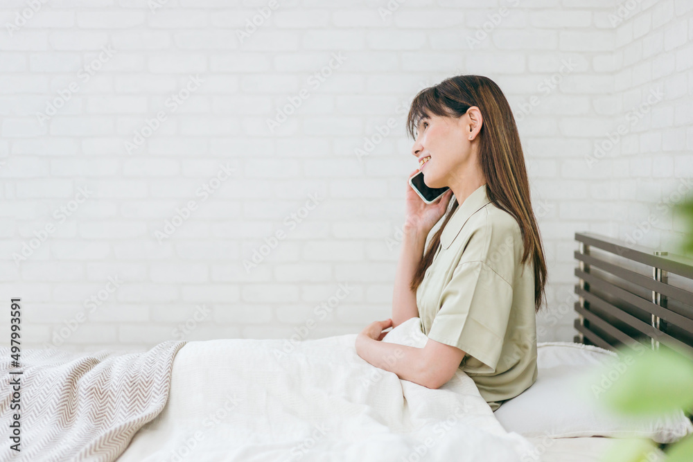 ベッドでスマホを使うミドル世代の女性(通話・笑顔)
ベッドでスマホを使うミドル世代の女性(通話・笑顔)
