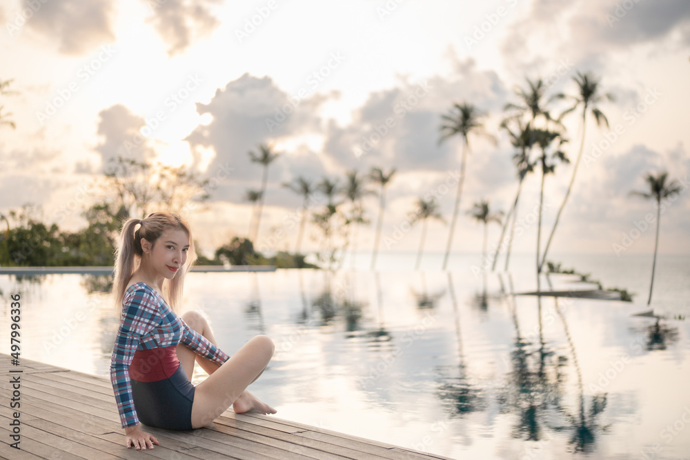 Woman in one piece swimsuit sitting near swimming pool, sunset moment.