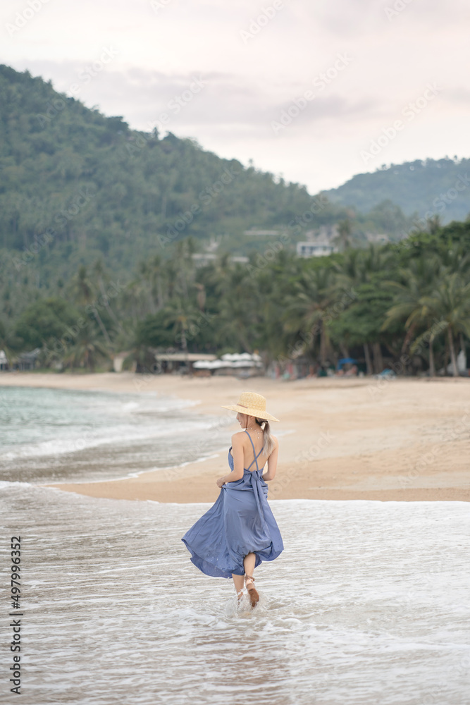 Back view of woman in blue dress and straw hat walk on the beach.