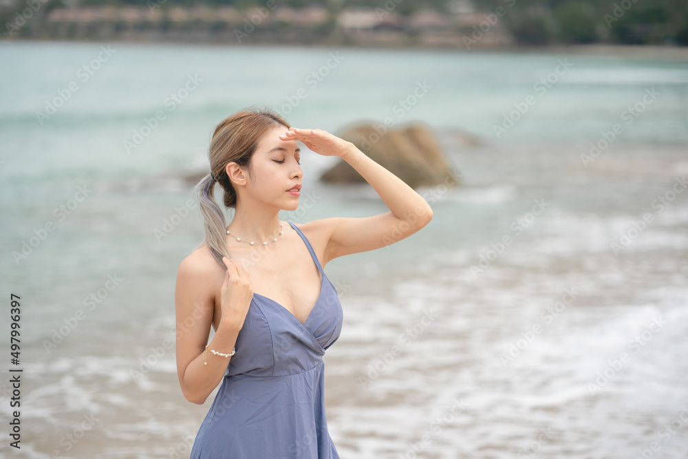 Woman in blue dress covering face by hand from sunlight on the beach.