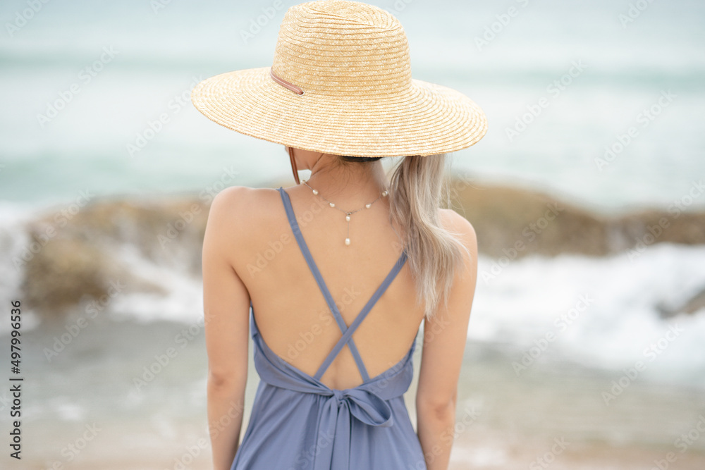 Back side of woman in  blue dress and straw hat, standing on a timber by the ocean.