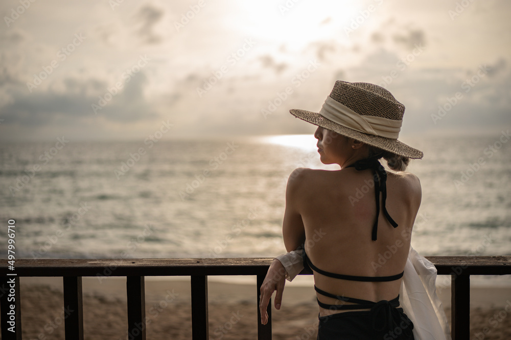 Back view of woman in black dress and straw hat standing on a balcony, ocean view.