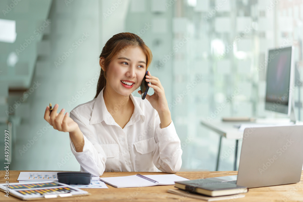 Smiling young Asian businesswoman sitting talking on the phone with laptop computer on desk in the office. Smiling young Asian businesswoman sitting talking on the phone with laptop computer on desk in the office.
