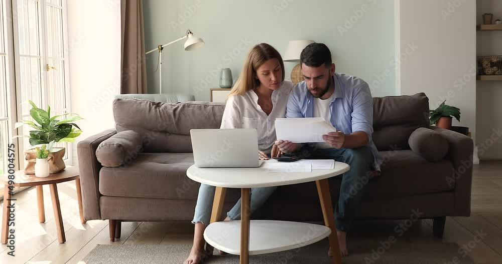 Millennial 35s married couple look at document, analyzing bank statement, examining domestic household rates, managing family budget, pay for mortgage loan bills, discuss expenses sit on couch at home Millennial 35s married couple look at document, analyzing bank statement, examining domestic household rates, managing family budget, pay for mortgage loan bills, discuss expenses sit on couch at home