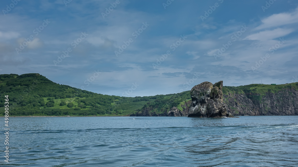 Kamchatka coast against the background of blue sky and ocean. Green hills and picturesque rocky cliffs. Ripples on the water. Avacha Bay. Kamchatka coast against the background of blue sky and ocean. Green hills and picturesque rocky cliffs. Ripples on the water. Avacha Bay.