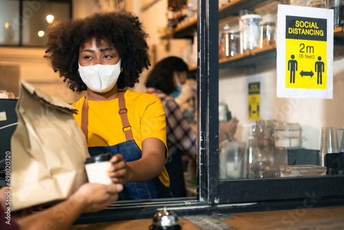 African Woman barista wear mask due to Covid-19 pandemic, receive drink on coffee bar counter. Restaurant worker giving takeaway delivery food bag.Small business social distance concept.