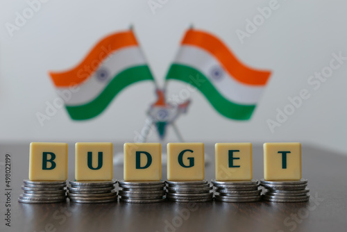 Letter blocks arranged as 'BUDGET' on pile of coins and blur Indian flag in the background.  Main focus is on letter blocks and coins. Flag is purposely blur.