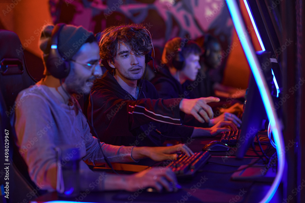 Obraz premium Curly-haired esports gamer pointing at computer monitor while helping Arabian rookie to complete level