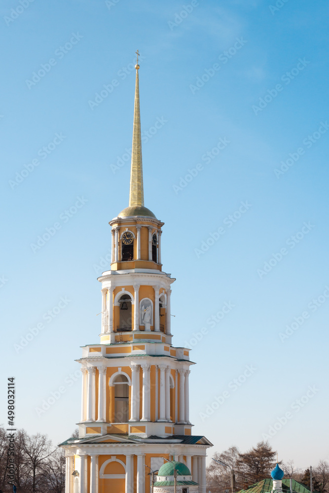 A orange and yellow golden orthodox church with a long pillar and a cross on top. A bell can be seen inside