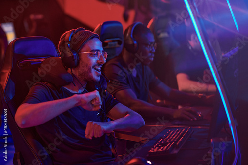 Happy young middle eastern professional gamer in headphones and eyeglasses dancing in front of computer while completing video game misson