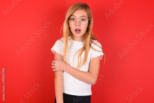 Shocked embarrassed blonde little kid girl wearing white t-shirt over red background keeps mouth widely opened. Hears unbelievable novelty stares in stupor photo