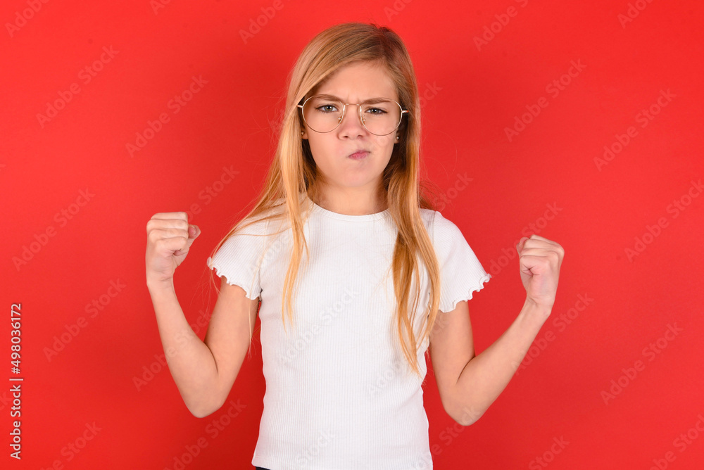 Fototapeta premium Irritated blonde little kid girl wearing white t-shirt over red background blows cheeks with anger and raises clenched fists expresses rage and aggressive emotions. Furious model