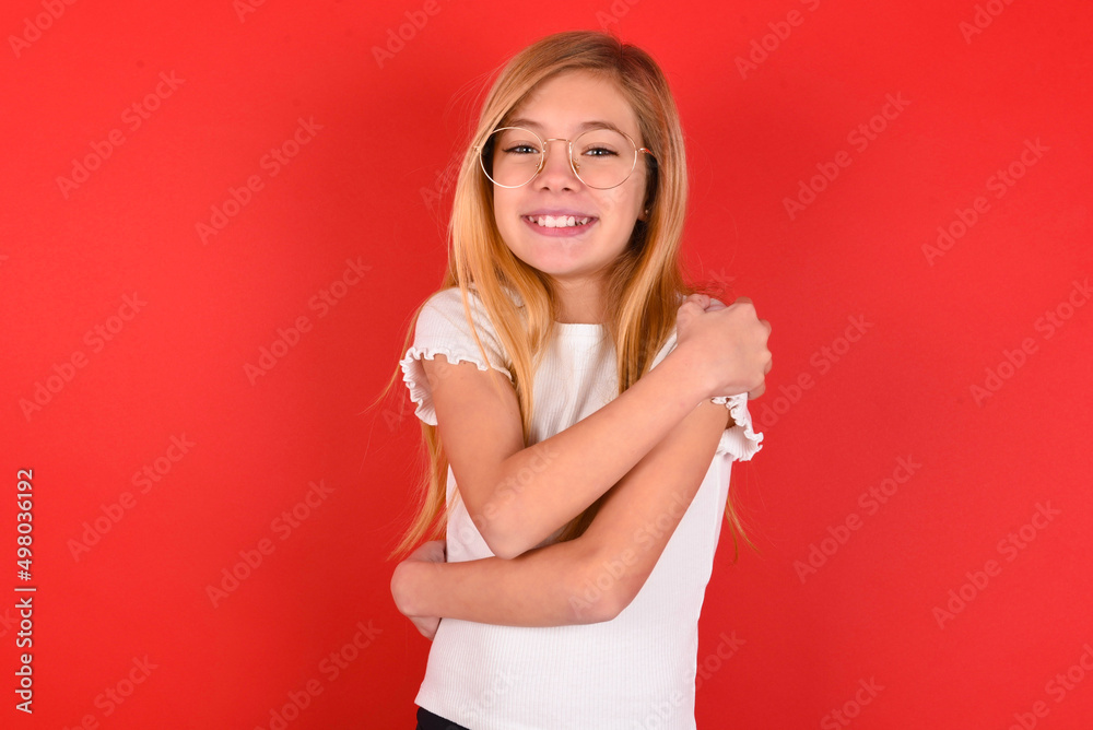Charming pleased blonde little kid girl wearing white t-shirt over red background embraces own body, pleasantly feels comfortable poses. Tenderness and self esteem concept Charming pleased blonde little kid girl wearing white t-shirt over red background embraces own body, pleasantly feels comfortable poses. Tenderness and self esteem concept