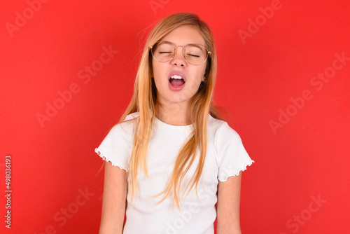 blonde little kid girl wearing white t-shirt over red background yawns with opened mouth stands. Daily morning routine
