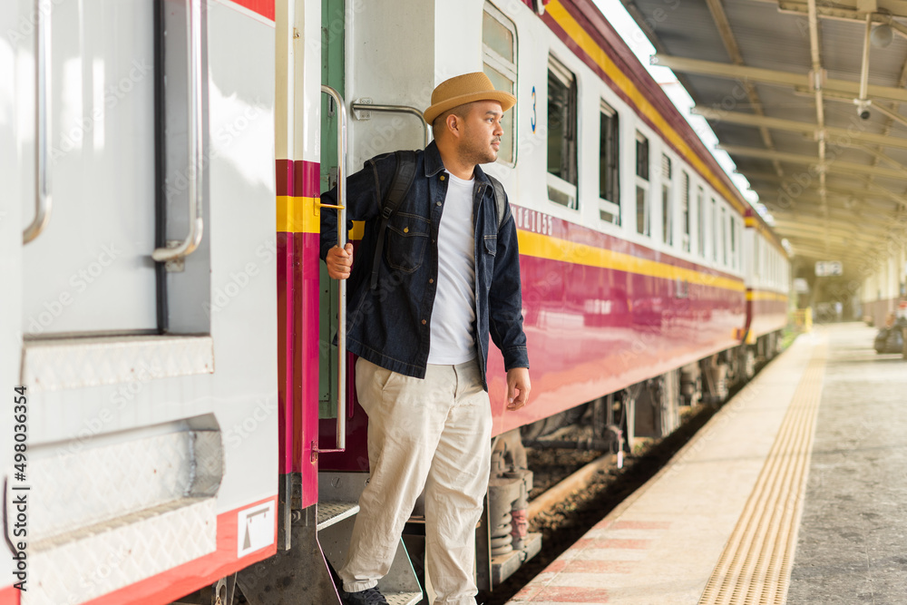 Young asian man traveler backpack in the train station. Backpacker male wear hat and denim jacket at the railway are going down the train ladder. Travel concept. The concept of a man traveling alone.