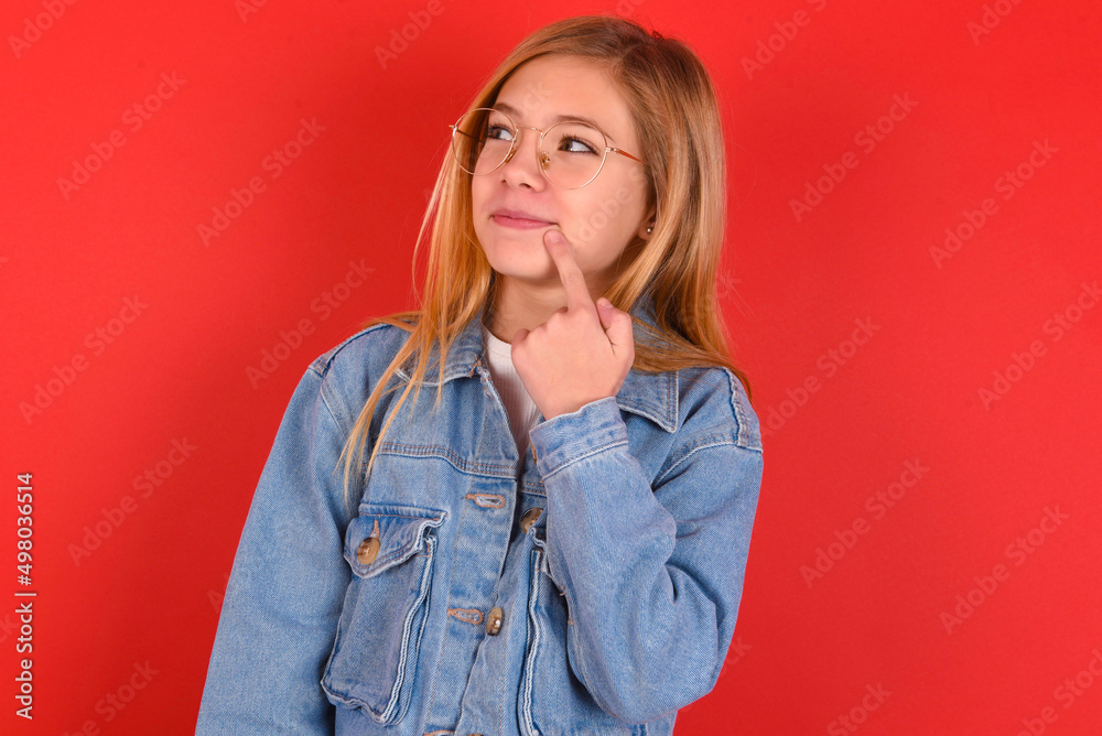 Lovely dreamy blonde little kid girl wearing denim jacket over red background keeps finger near lips looks aside copy space. Lovely dreamy blonde little kid girl wearing denim jacket over red background keeps finger near lips looks aside copy space.
