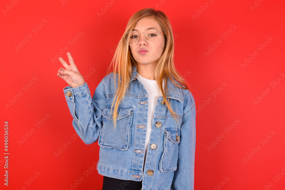 blonde little kid girl wearing denim jacket over red background makes peace gesture keeps lips folded shows v sign. Body language concept blonde little kid girl wearing denim jacket over red background makes peace gesture keeps lips folded shows v sign. Body language concept