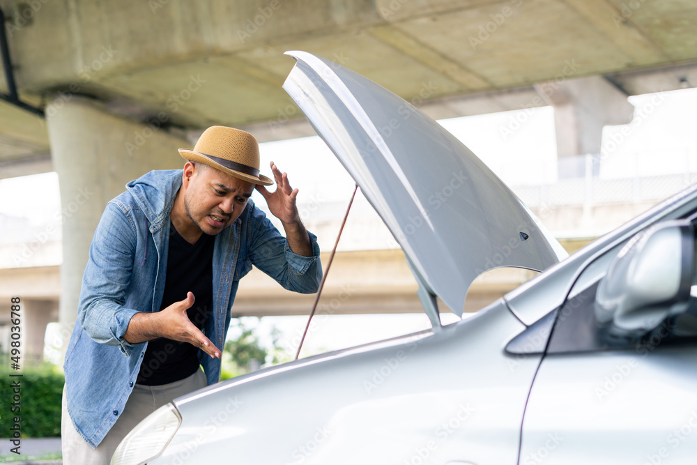 Frustrated young asian man looking at broken down car engine on street waiting for assistance to arrive and recover or fix the vehicle. Male angry stand front a broken car. Broken car while traveling. Frustrated young asian man looking at broken down car engine on street waiting for assistance to arrive and recover or fix the vehicle. Male angry stand front a broken car. Broken car while traveling.