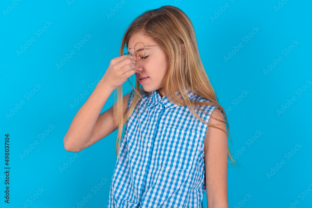 Very upset, little kid girl with glasses wearing plaid shirt over blue background touching nose between closed eyes, wants to cry, having stressful relationship or having troubles with work Very upset, little kid girl with glasses wearing plaid shirt over blue background touching nose between closed eyes, wants to cry, having stressful relationship or having troubles with work