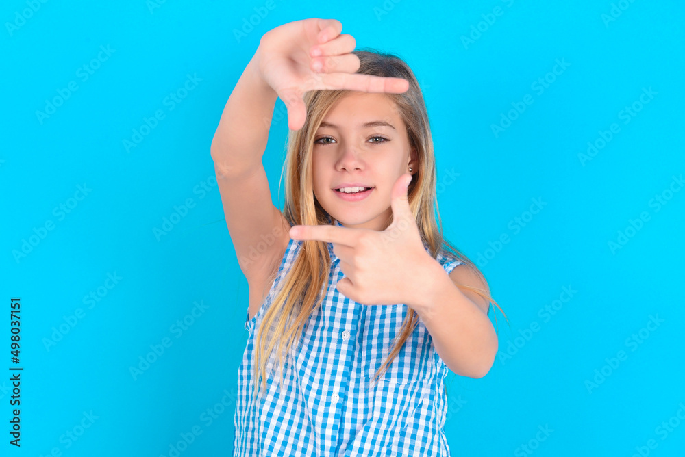 Positive little kid girl with glasses wearing plaid shirt over blue background  with cheerful expression, has good mood, gestures finger frame actively at camera.