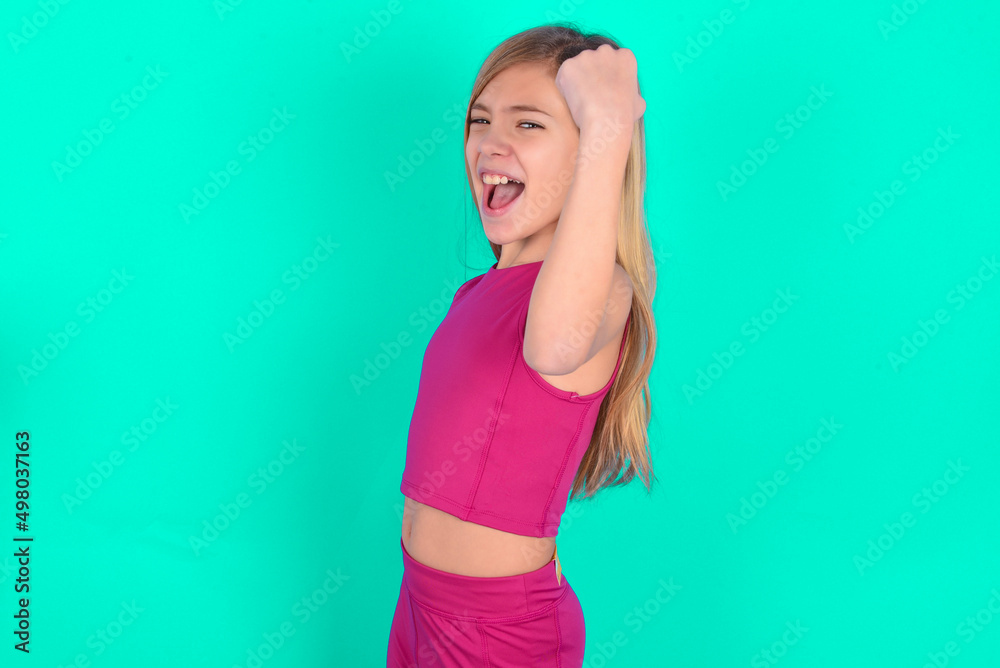 Overjoyed blonde little kid girl wearing pink sport clothes over green background glad to receive good news, clenching fist and making winning gesture. Overjoyed blonde little kid girl wearing pink sport clothes over green background glad to receive good news, clenching fist and making winning gesture.
