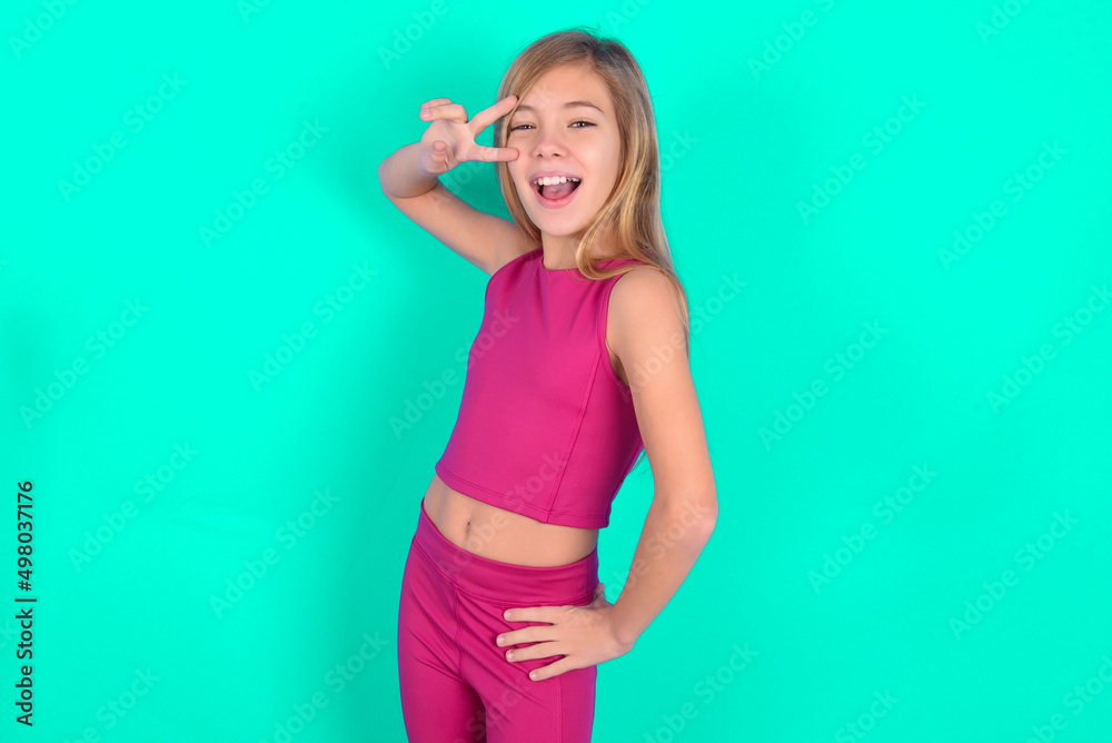 blonde little kid girl wearing pink sport clothes over green background making v-sign near eyes. Leisure, coquettish, celebration, and flirt concept. blonde little kid girl wearing pink sport clothes over green background making v-sign near eyes. Leisure, coquettish, celebration, and flirt concept.