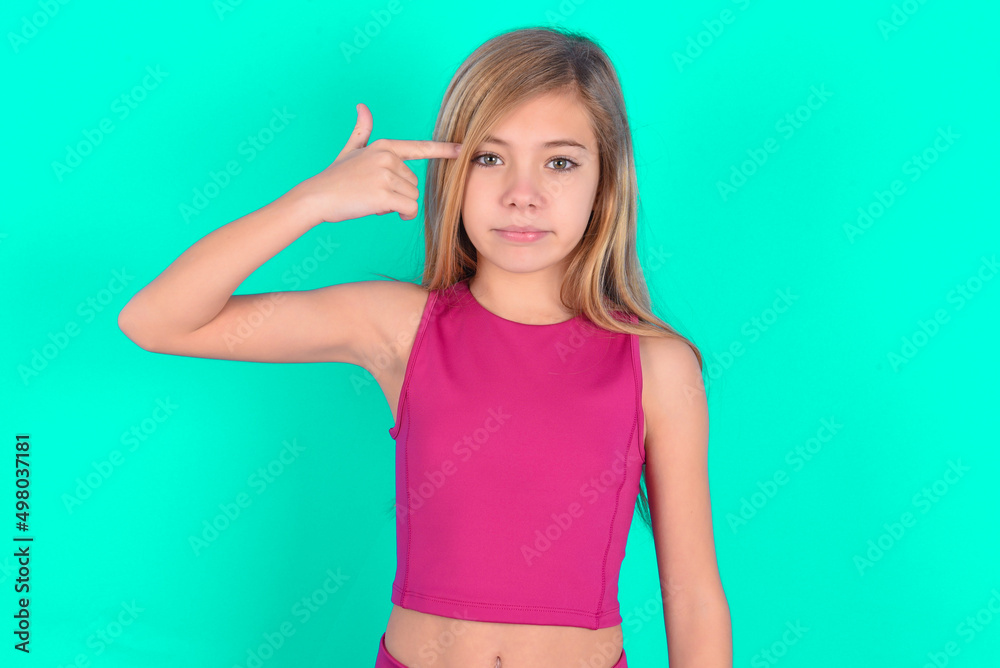 Unhappy blonde little kid girl wearing pink sport clothes over green background curves lips and makes suicide gesture, shoots in temple with hand, tries kill himself. Unhappy blonde little kid girl wearing pink sport clothes over green background curves lips and makes suicide gesture, shoots in temple with hand, tries kill himself.