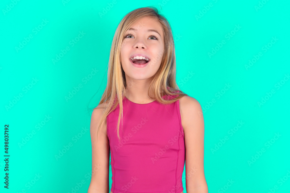 Surprised blonde little kid girl wearing pink sport clothes over green background , shrugs shoulders, looking sideways, being happy and excited. Sudden reactions concept. Surprised blonde little kid girl wearing pink sport clothes over green background , shrugs shoulders, looking sideways, being happy and excited. Sudden reactions concept.