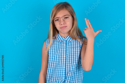 Unhappy little kid girl with glasses wearing plaid shirt over blue background imitates gun shoot makes suicide gesture keeps two fingers on temples.