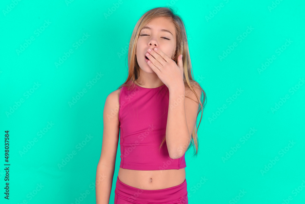 Sleepy blonde little kid girl wearing pink sport clothes over green background yawning with messy hair, feeling tired after sleepless night, yawning, covering mouth with palm. Sleepy blonde little kid girl wearing pink sport clothes over green background yawning with messy hair, feeling tired after sleepless night, yawning, covering mouth with palm.
