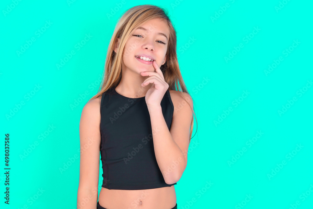 Carefree successful blonde little kid girl wearing black sport clothes over green background touching jawline gazing camera tilting head grinning white teeth delighted. Dental care concept.