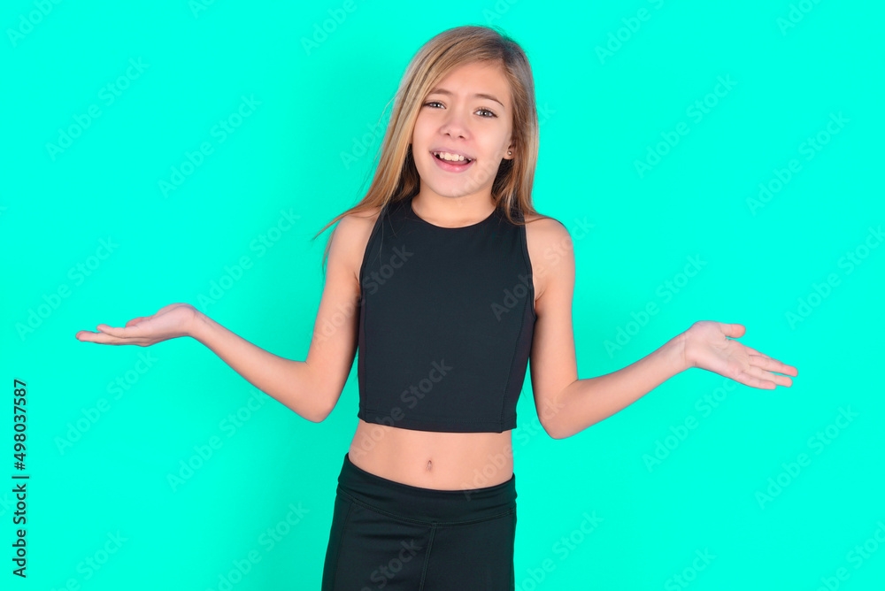 So what? Portrait of arrogant blonde little kid girl wearing black sport clothes over green background shrugging hands sideways smiling gasping indifferent, telling something obvious. So what? Portrait of arrogant blonde little kid girl wearing black sport clothes over green background shrugging hands sideways smiling gasping indifferent, telling something obvious.