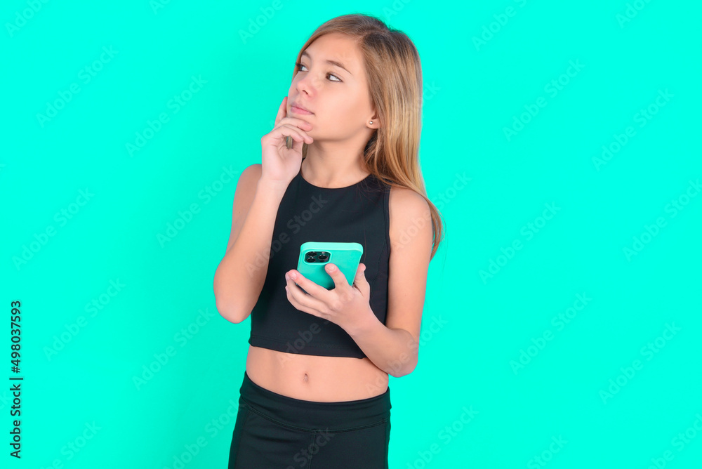 Thinking dreaming blonde little kid girl wearing black sport clothes over green background using mobile phone and holding hand on face. Taking decisions and social media concept. Thinking dreaming blonde little kid girl wearing black sport clothes over green background using mobile phone and holding hand on face. Taking decisions and social media concept.