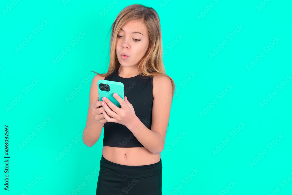 blonde little kid girl wearing black sport clothes over green background looks with bugged eyes, holds modern smart phone, receives unexpected message from friend, reads reminder. blonde little kid girl wearing black sport clothes over green background looks with bugged eyes, holds modern smart phone, receives unexpected message from friend, reads reminder.