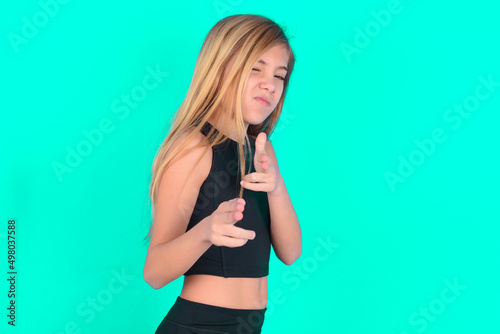 Hey you, bang. Joyful and charismatic good-looking blonde little kid girl wearing black sport clothes over green background winking and pointing with finger pistols at camera happily and cheeky.