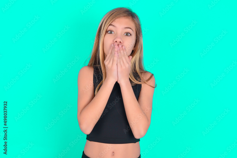 blonde little kid girl wearing black sport clothes over green background keeps hands on mouth, looks with eyes full of disbelief, being puzzled with amount of work blonde little kid girl wearing black sport clothes over green background keeps hands on mouth, looks with eyes full of disbelief, being puzzled with amount of work