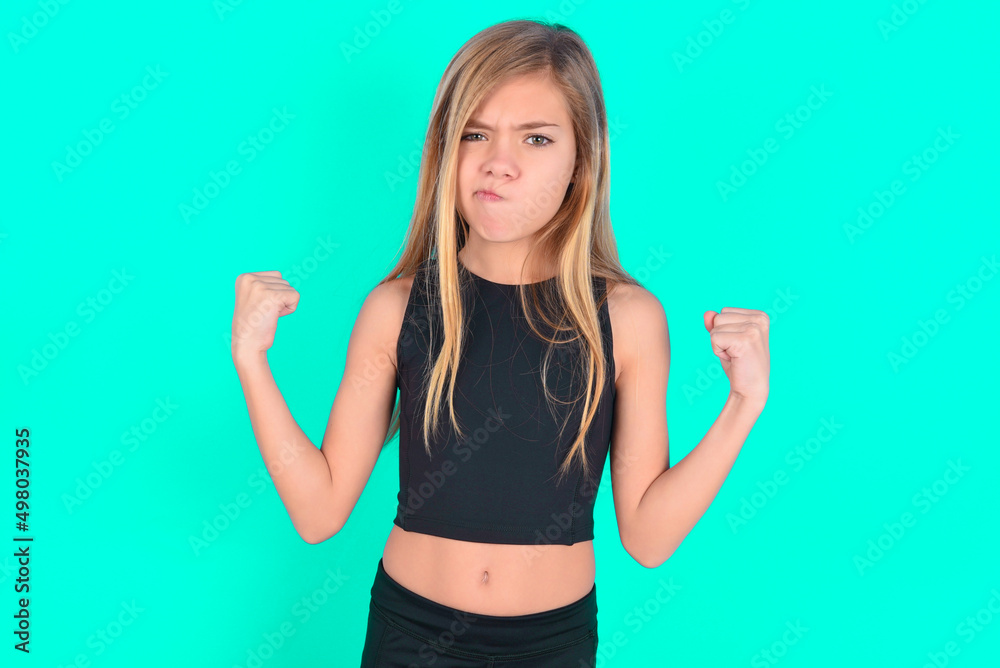 Irritated blonde little kid girl wearing black sport clothes over green background blows cheeks with anger and raises clenched fists expresses rage and aggressive emotions. Furious model Irritated blonde little kid girl wearing black sport clothes over green background blows cheeks with anger and raises clenched fists expresses rage and aggressive emotions. Furious model