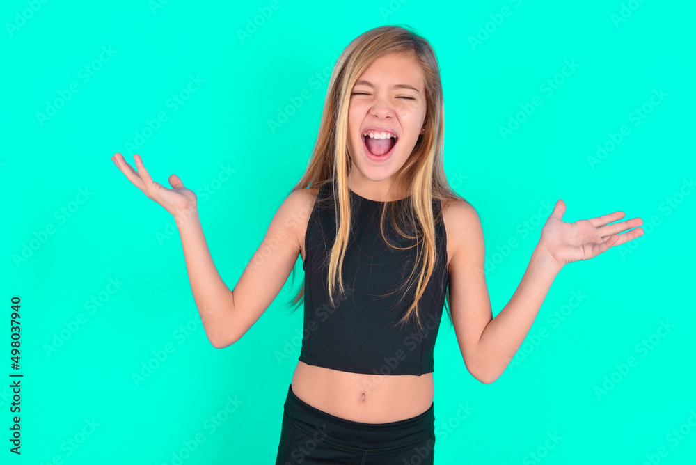 Fototapeta premium Crazy outraged blonde little kid girl wearing black sport clothes over green background screams loudly and gestures angrily yells furiously. Negative human emotions feelings concept