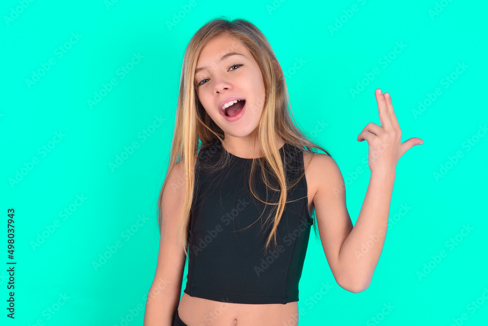 blonde little kid girl wearing black sport clothes over green background foolishness around shoots in temple with fingers makes suicide gesture. Funny model makes finger gun pistol blonde little kid girl wearing black sport clothes over green background foolishness around shoots in temple with fingers makes suicide gesture. Funny model makes finger gun pistol