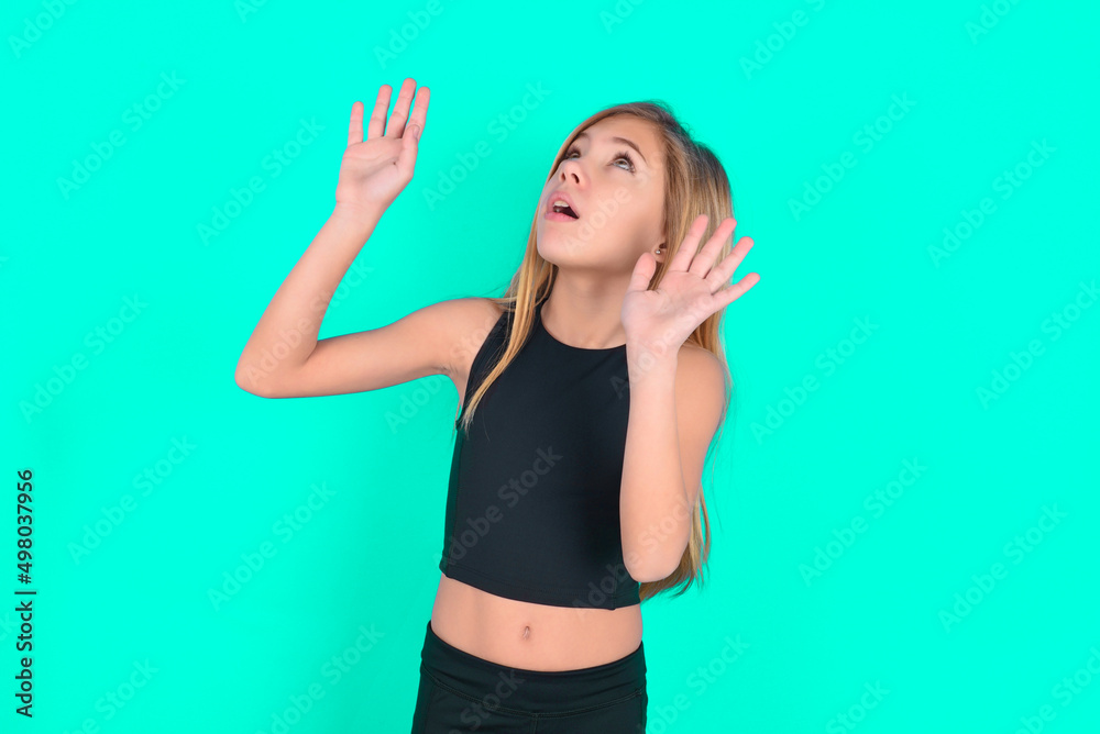 blonde little kid girl wearing black sport clothes over green background keeps palms forward and looks with fright above on ceiling tries to defense herself from invisible danger opens mouth. blonde little kid girl wearing black sport clothes over green background keeps palms forward and looks with fright above on ceiling tries to defense herself from invisible danger opens mouth.