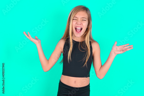 Crazy outraged blonde little kid girl wearing black sport clothes over green background screams loudly and gestures angrily yells furiously. Negative human emotions feelings concept