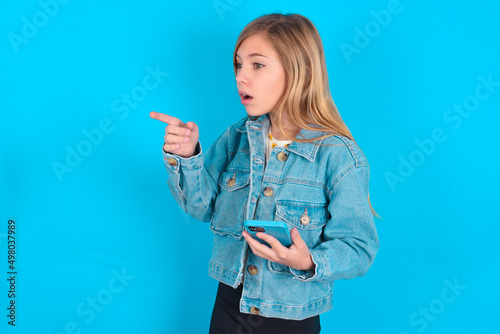 Stunned blonde little kid girl wearing denim jacket over blue background points sideways right copy space, recommends product, sees astonishing thing photo