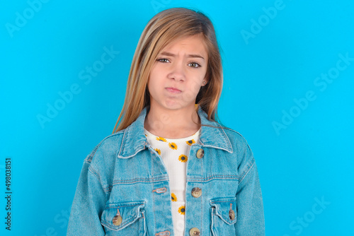 Cuadro en lienzo Displeased blonde little kid girl wearing denim jacket over blue background frowns face feels unhappy has some problems