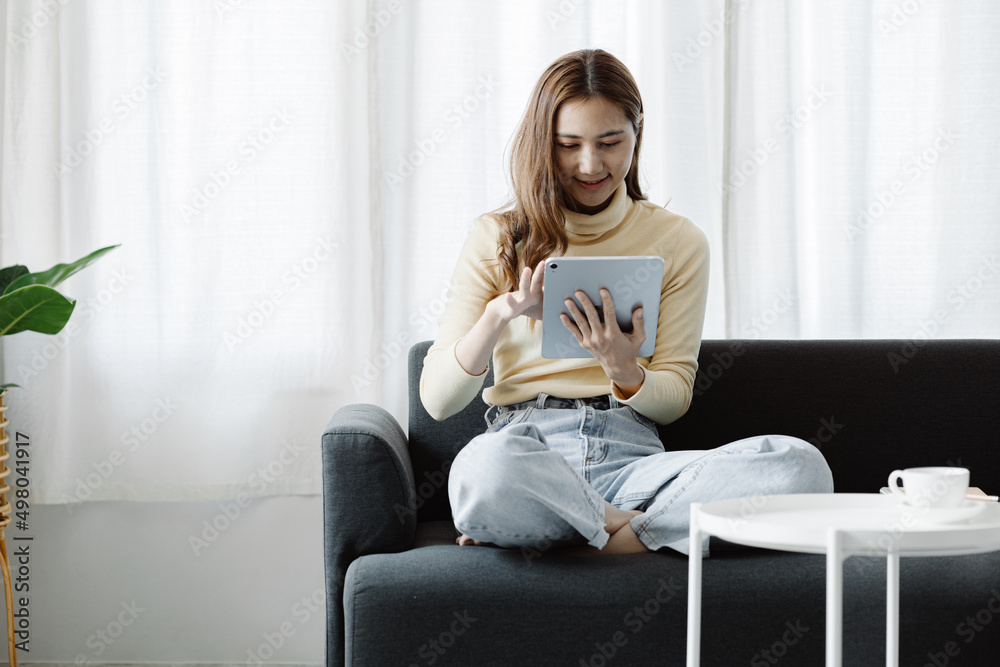 A woman plays a tablet on the sofa in her living room at home, she is resting on weekends after a hard day's work, she relaxes by watching movies and listening to music on her tablet. Holiday concept. A woman plays a tablet on the sofa in her living room at home, she is resting on weekends after a hard day's work, she relaxes by watching movies and listening to music on her tablet. Holiday concept.