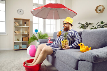 Funny man spends summer vacation at home and sitting on sofa pretending he is on beach. Man drinking cocktail and keeping his feet in plastic bowl sitting under beach umbrella in living room.
