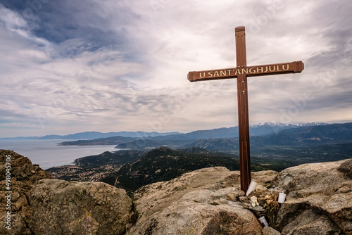 Cross at the summit of Cima Sant'Anghjulu near Corbara in the Balagne region of Corsica with the coastline and snow capped mountains in the distance