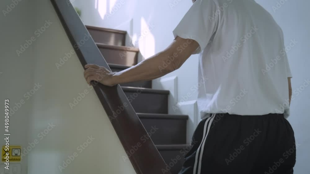 Asian man wearing glasses with short hair, retired, 70 years old, resting at home Walk slowly up the stairs of the house with caution holding onto the railing. for safety Asian man wearing glasses with short hair, retired, 70 years old, resting at home Walk slowly up the stairs of the house with caution holding onto the railing. for safety
