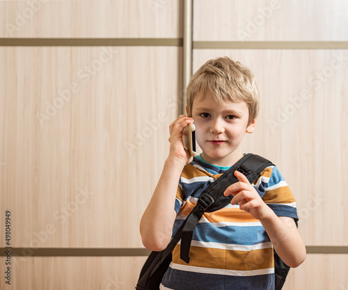 a little boy in a striped T-shirt and with a backpack on his back is watching, calling on a smartphone, which he holds in his hand and with the other pointing a finger at the photographer.