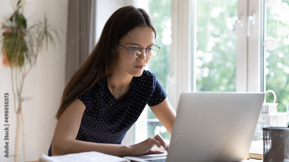 Busy student girl in glasses studying use laptop, prepare for university exams looks focused, serious concentrated woman working sit at table staring at computer screen. Modern tech, e-learn concept
