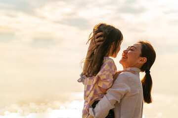 Happy Asian family on beach vacation. Mother carrying and kissing  little daughter while walking on tropical beach at summer sunset. Mom and child girl kid enjoy and fun outdoor lifestyle on the beach
